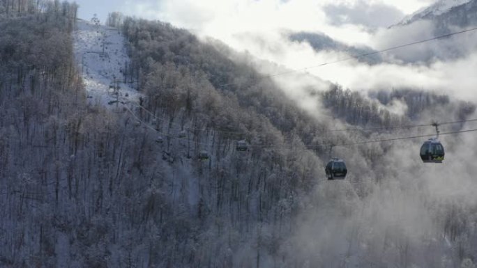 在薄雾笼罩的雪山之间移动的滑雪索道遵循鸟瞰图。滑雪电梯将人们运送到多云的山背景上的雪坡。无人机上方的