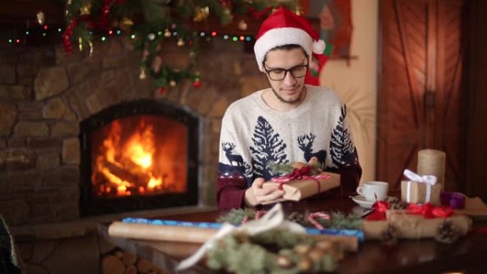 大胡子的男人坐在壁炉附近的礼物上系弓。戴着圣诞帽的家伙用纸包装盒子，用冷杉树枝，圆锥体，甘蔗糖果装饰