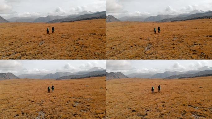 一对带着帽子和墨镜的大背包的旅行者男人和女人的鸟瞰图沿着被史诗般的山脉环绕的高山高原行走