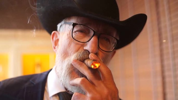 穿着宽边帽的时尚老人和丰富的蓝色男士套装室内吸烟雪茄