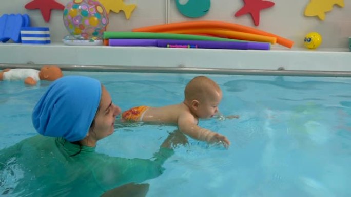 母亲用手支撑着婴儿，这只手漂浮在儿童游泳池的水中。婴儿游到游泳池边，环顾四周，微笑着。婴儿游泳。特写