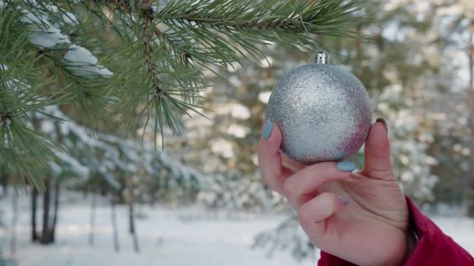 关闭女性手触摸挂在户外圣诞树上的银球。年轻女子的手在白雪皑皑的森林中的新年树上挂球玩具。
