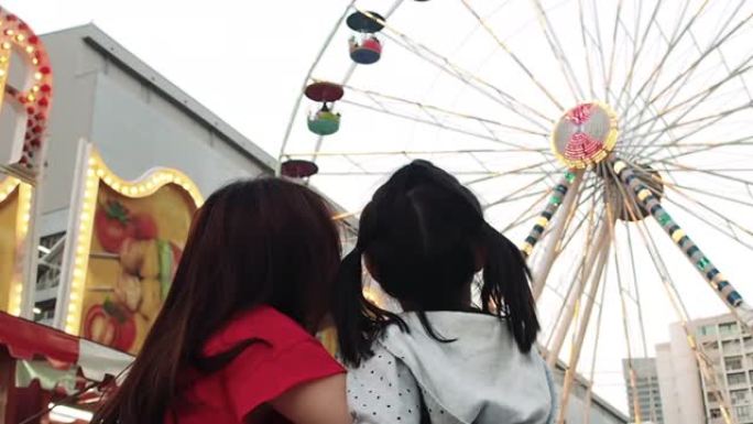 享受游乐场的概念。亚洲母亲和女儿感到快乐，并以摩天轮背景指向。