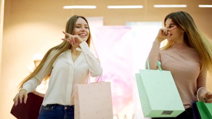 两个带着购物袋的快乐年轻女性在商场橱窗展示附近慢动作跳舞，庆祝购物愉快
