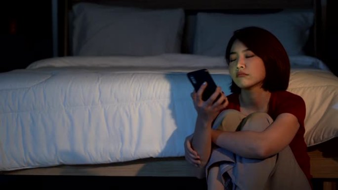 4k沮丧的年轻美丽的亚洲女人独自坐在卧室地板上，使用智能手机。不快乐的压力女人十几岁的女孩坐在地板上