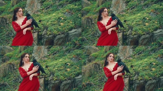 森林豪华的仙女与乌鸦在她的手臂。迷人的女人在红色豪华长裙与深领口拖曳抚摸鸟与温柔。绿草和石头。野生动