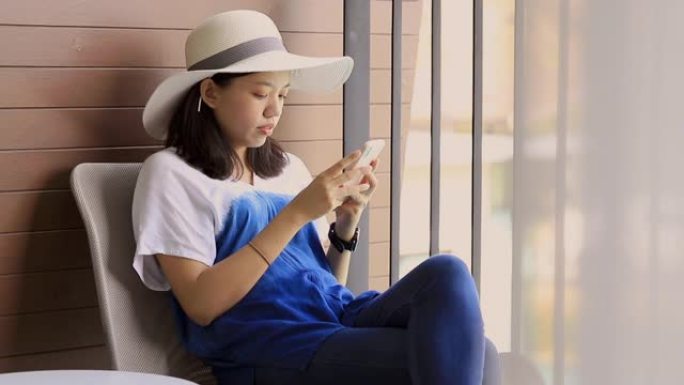 年轻漂亮的亚洲女人戴着宽帽子，使用智能手机坐在酒店房间阳台的椅子上。假日旅行概念。慢动作