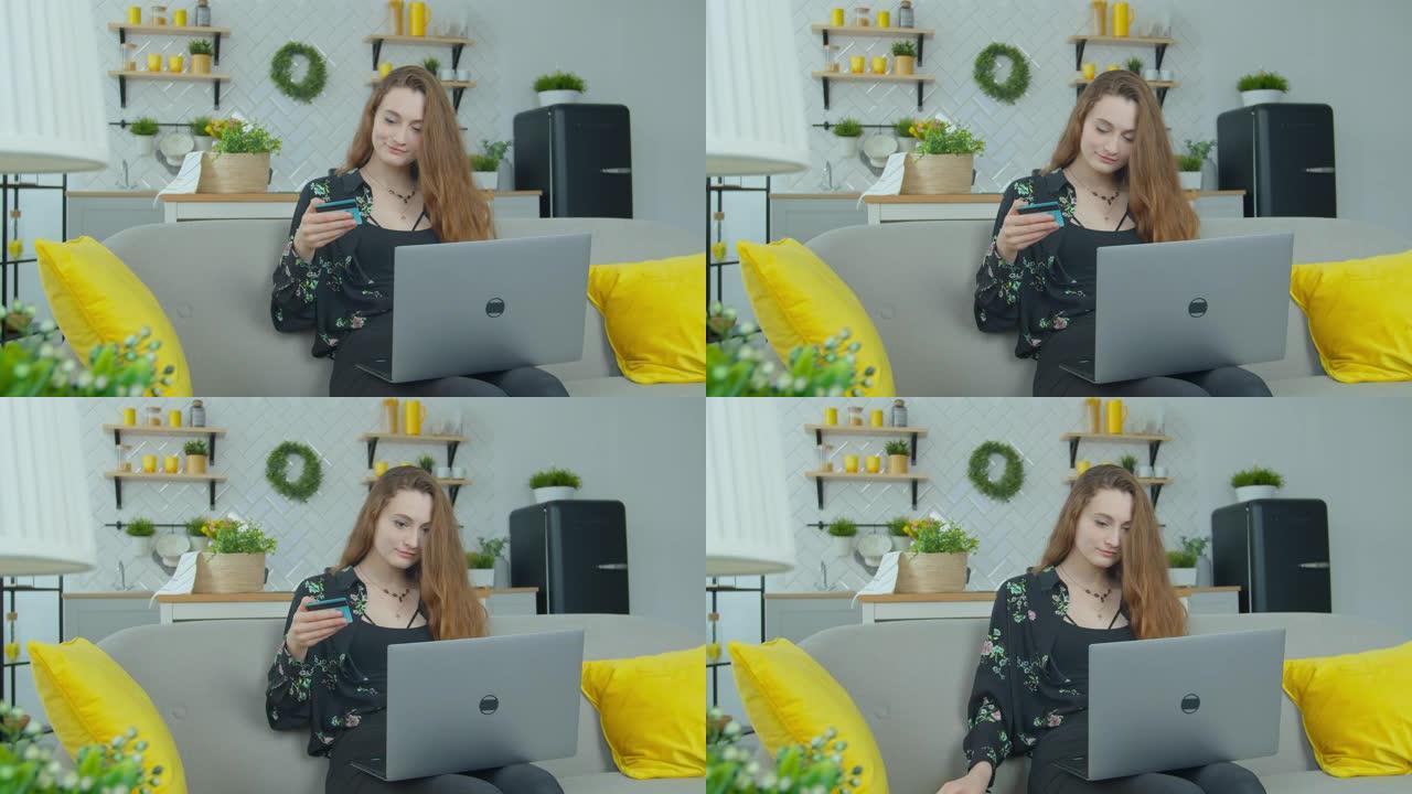 女孩正在用现代笔记本电脑在家坐着微笑着拿着银行卡进行在线支付。
