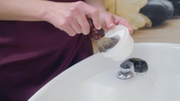 造型师用剃须刷准备剃须泡沫。理发师的手靠近。模糊背景下美发沙龙的盥洗台。