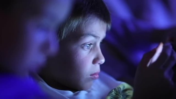 平板电脑屏幕前的小男孩，蓝光发光的孩子在黑暗中看着智能手机屏幕