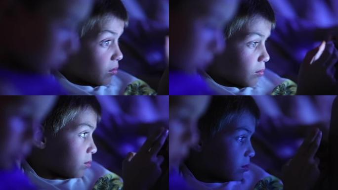 平板电脑屏幕前的小男孩，蓝光发光的孩子在黑暗中看着智能手机屏幕