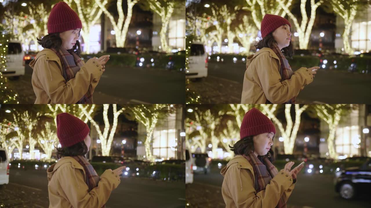 年轻的亚洲妇女戴上羊毛帽子和围巾，晚上在城市的智能手机上请求并等待Uber。
