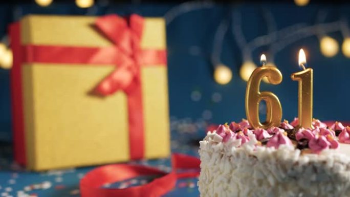 白色生日蛋糕编号61点灯燃烧的金色蜡烛，带灯的蓝色背景和用红丝带绑起来的礼物黄色盒子。特写
