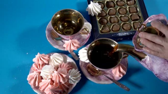 红茶被倒入蓝色背景上镀金的粉红色瓷杯中。旁边是棉花糖和巧克力的碟子。从顶部看。特写。4K