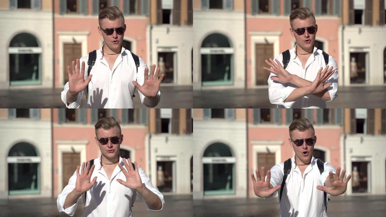 年轻恼怒的男人留在街上并在相机上打手势的真实面部情绪。男子显示停止或没有举手和交叉的标志。负面情绪的