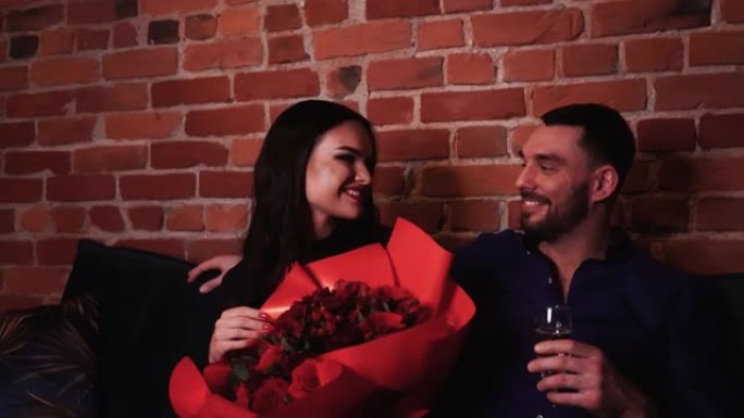 相爱的美丽年轻夫妇互相享受，在沙发上放着鲜花和香槟。用蜡烛在浪漫的气氛中微笑。