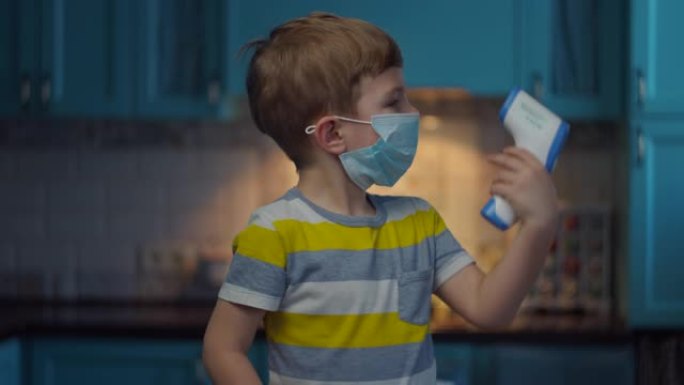 脸上戴着医用面具的金发男孩用红外温度计看着相机测量温度。孩子在家里的医疗面具在病毒的自我隔离。停止流