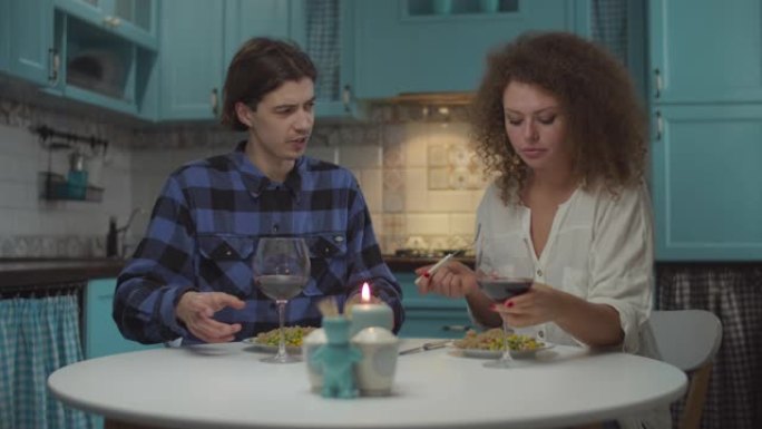 年轻快乐的20多岁夫妇穿着休闲服装，在舒适的蓝色厨房享用晚餐时喝红酒。男人和女人在慢动作中在家吃饭和