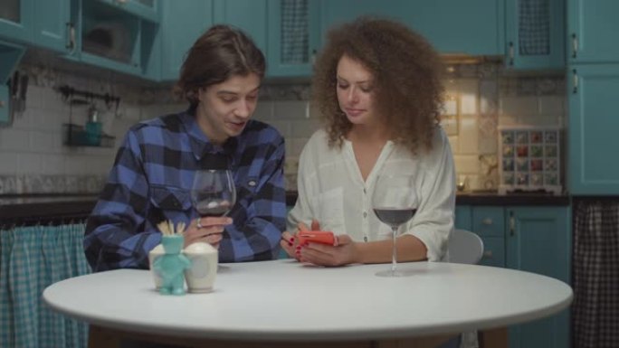 20多岁的年轻夫妇在蓝色厨房喝带小工具的红酒。男人和女人在家里慢动作喝酒时，兴致地看着女人手中的手机