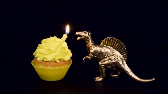 金色恐龙在黑色背景上隔离的亮黄色纸杯蛋糕上吹出蜡烛。生日快乐概念。高清视频。生日蛋糕和玩具恐龙