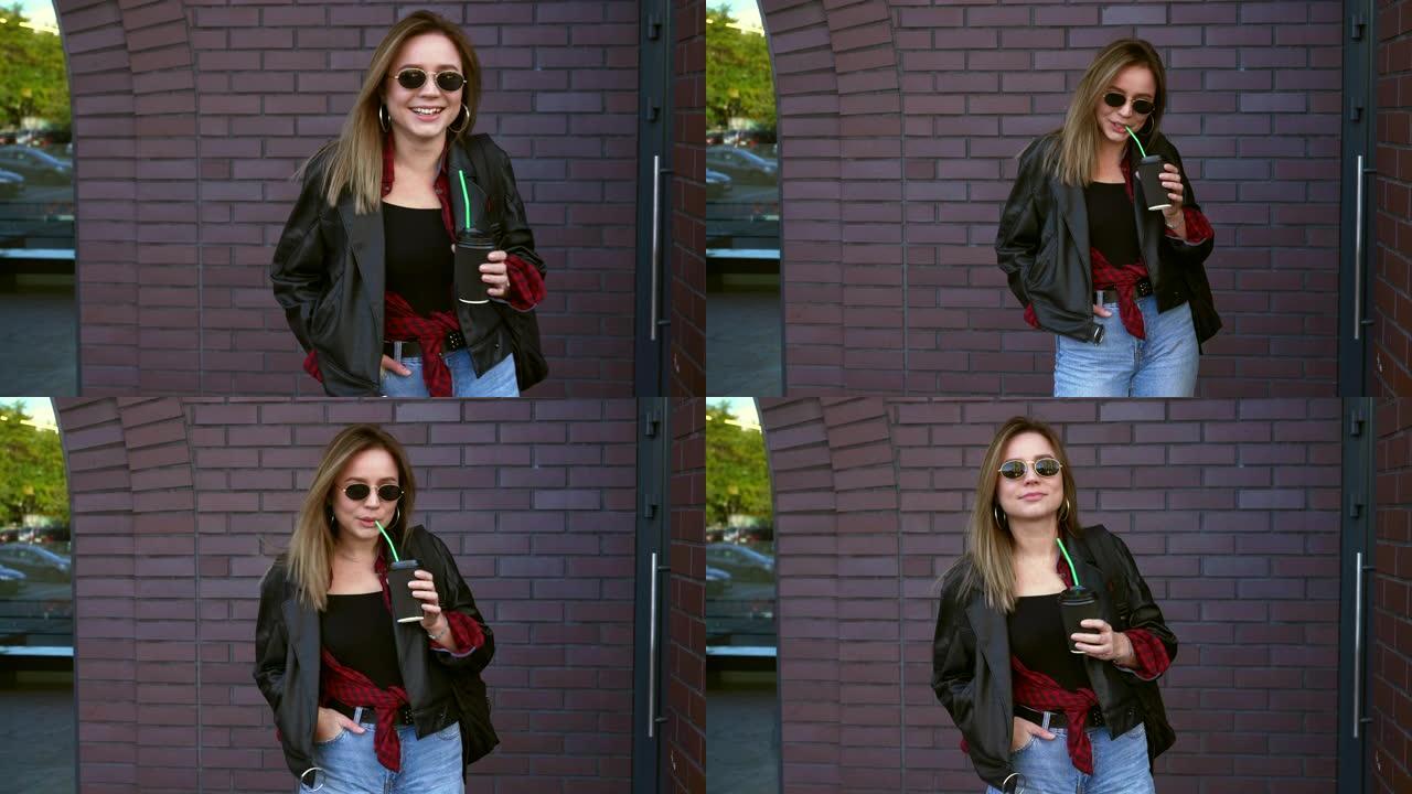 年轻漂亮的女学生站在秋天的砖墙附近，戴着墨镜，手里拿着咖啡，笑着。