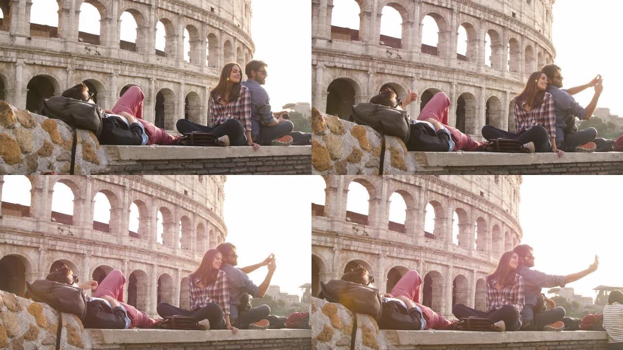 三个年轻的朋友游客日落时坐在罗马斗兽场前用智能手机自拍带背包太阳镜快乐美丽的女孩长发