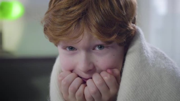 害怕的高加索红发男孩的特写镜头，坐在室内盖着毯子，环顾四周。可爱的小孩独自离开了家。童年，恐惧，生活