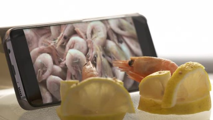 两只冷冻大虾坐在柠檬椅上观看手机屏幕上的煮虾食谱视频。