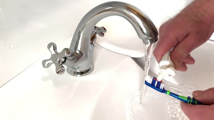 男人挤压小管，将牙膏放在刷子上，将其放在水槽上，水从水龙头流出。用刷子和牙膏管近距离观察人的手