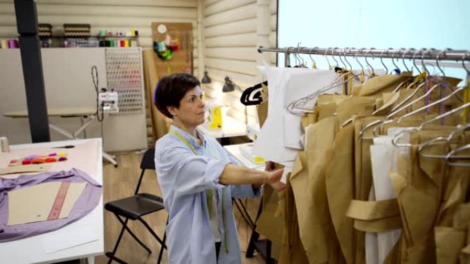 追踪中年女裁缝中拍，用布和纸缝制图案，站立并浏览缝纫工作室挂在铁轨上的图案