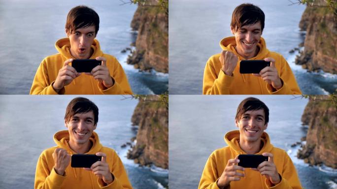 庆祝男子旅行者在户外的智能手机应用程序上在海浪上方的悬崖上获胜。在旅行中欢呼雀跃的胜利者庆祝好消息或