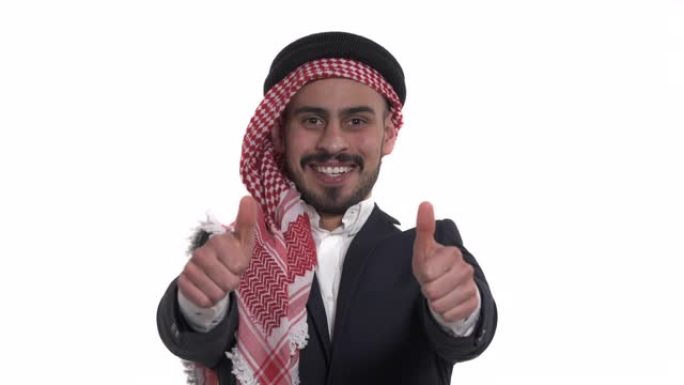 穿着keffiyeh的年轻英俊的阿拉伯男子通过竖起大拇指来表达幸福 (白色孤立)。真实的情感。肢体语