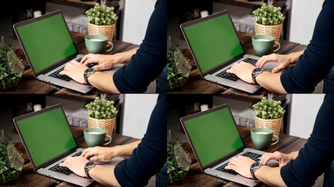桌子上的人正在绿色的笔记本电脑屏幕上浏览互联网。在光线充足、舒适的公寓里。一名男子在阁楼办公室工作