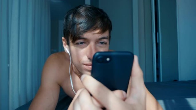 英俊的裸笑男人戴着耳机躺在家里的床上刷手机