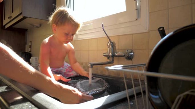 孩子在厨房里帮助母亲洗杯子和盘子。蹒跚学步的孩子坐在水槽里帮助父母。家务背景。职责和孩子。儿科推荐。