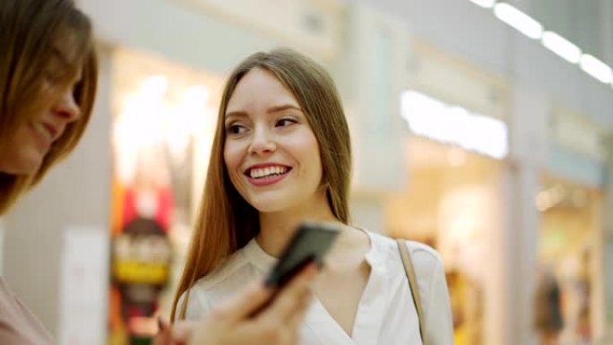 两个年轻漂亮的女性朋友带着购物袋在智能手机上浏览图片，快乐地聊天站在购物中心，放大平移镜头