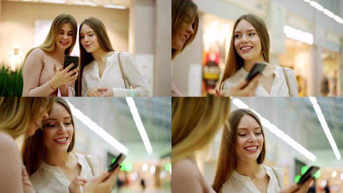 两个年轻漂亮的女性朋友带着购物袋在智能手机上浏览图片，快乐地聊天站在购物中心，放大平移镜头