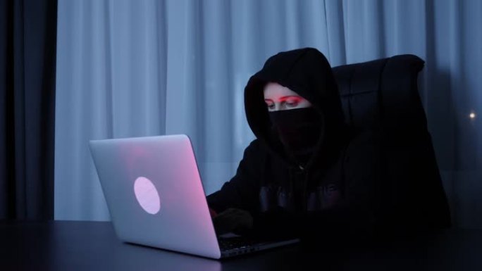 女黑客坐在黑暗的房间里，在笔记本电脑键盘上打字。女人在屏幕上戴着黑色面具和引擎盖编码，并打破密码。专