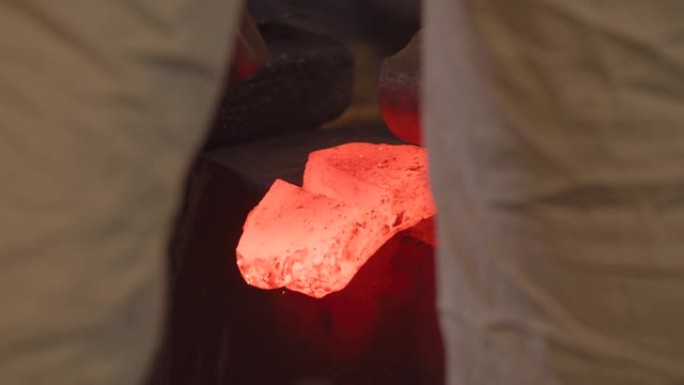 黑色史密斯大师的两腿之间的视图用大锤敲打红色的热锭，以折叠钢材并形成哈蒙。传统的日本锻造方法。日本工
