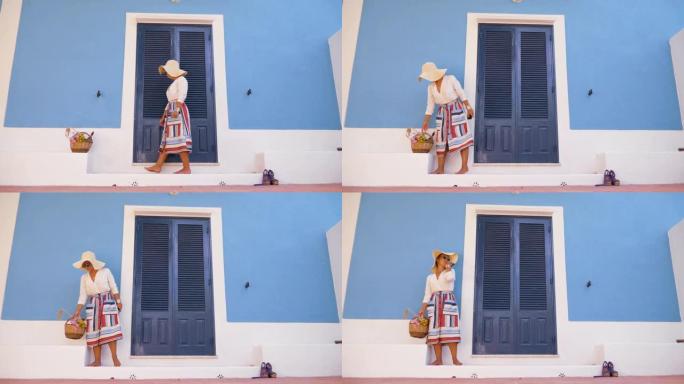 年轻幸福的女人站在带花篮的蓝屋入口露台的台阶上。时尚白衬衫，大帽子，彩色裙子和太阳镜。意大利蓬扎岛。