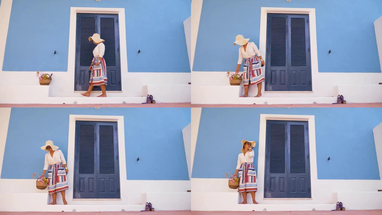 年轻幸福的女人站在带花篮的蓝屋入口露台的台阶上。时尚白衬衫，大帽子，彩色裙子和太阳镜。意大利蓬扎岛。