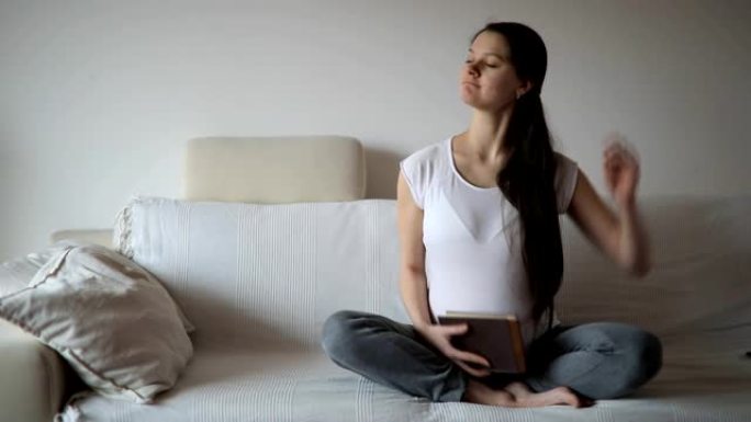 年轻的孕妇坐在家里房间的白色沙发上，停下来看书，然后伸展身体。