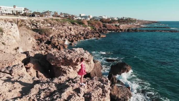 穿着红色连衣裙的女孩在风中拍打着，站在岩石悬崖上，双手分开，强烈的海浪撞击海岸，飞溅并产生泡沫。穿着