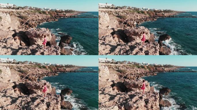 穿着红色连衣裙的女孩在风中拍打着，站在岩石悬崖上，双手分开，强烈的海浪撞击海岸，飞溅并产生泡沫。穿着