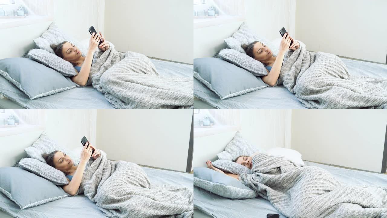 年轻迷人的女人醒来时拿着智能手机浏览社交媒体，放下手机，在床上享受早晨。