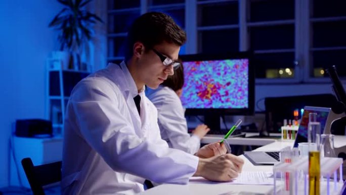 年轻的男性科学家检查试管中的液体，写下测试结果并使用笔记本电脑，而他的女同事在生物化学实验室的计算机