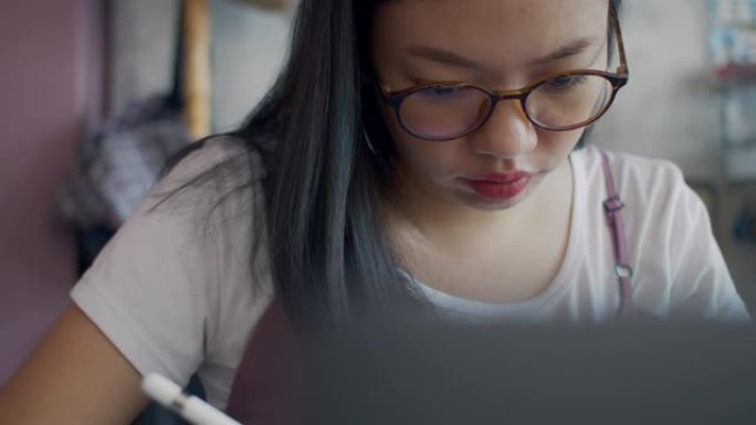 亚洲女商人戴着眼镜思考，在家庭办公室工作，用平板电脑、智能手机和笔记本电脑。写字和画画决心专注于工作