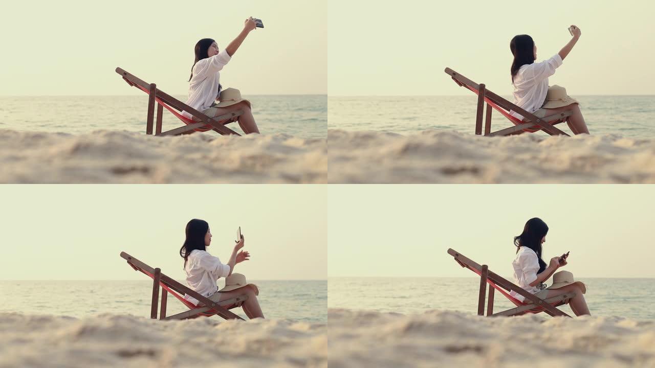 快乐的亚洲女人感到轻松躺在沙滩上的躺椅上，并使用智能手机自拍上传到社交媒体交流。假期暑期概念。