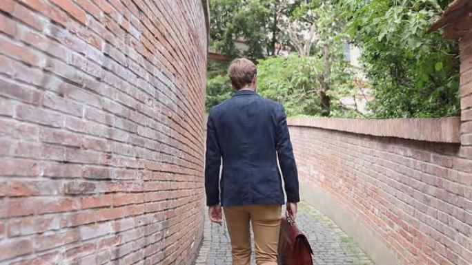 一个年轻潇洒随意的男人走在他的手提箱靠近砖墙的背影