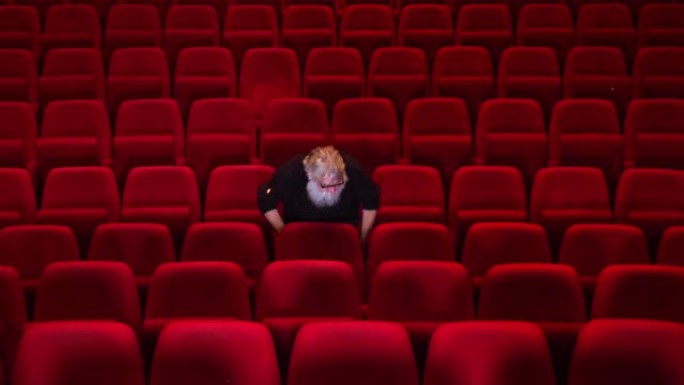 一个留着白胡子的男人坐在空荡荡的电影院或剧院里，进入